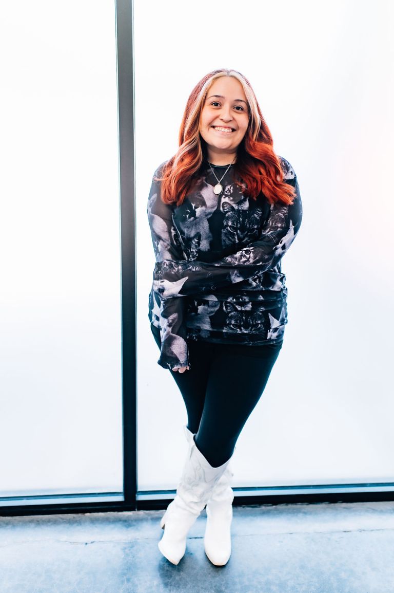 Office Manager Danielly standing confidently in a cat-patterned top and white boots, smiling against a bright background, representing Veterinary Vision Specialists' commitment to compassionate animal care.